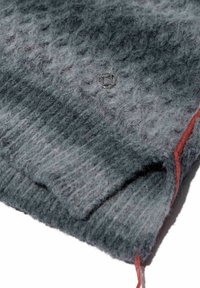 Grauer Wollmischungspullover mit ribbeligem Saum, der eine glatte, flauschige Textur aufweist und ein kleines silbernes Emblem hat. Ein Akzent aus rotem Stick entsteht sichtbar.