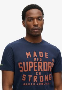 Camiseta de algodón azul marino con un gran texto naranja que dice "HECHO SUPERDRY FUERTE", con cuello redondo y mangas cortas.