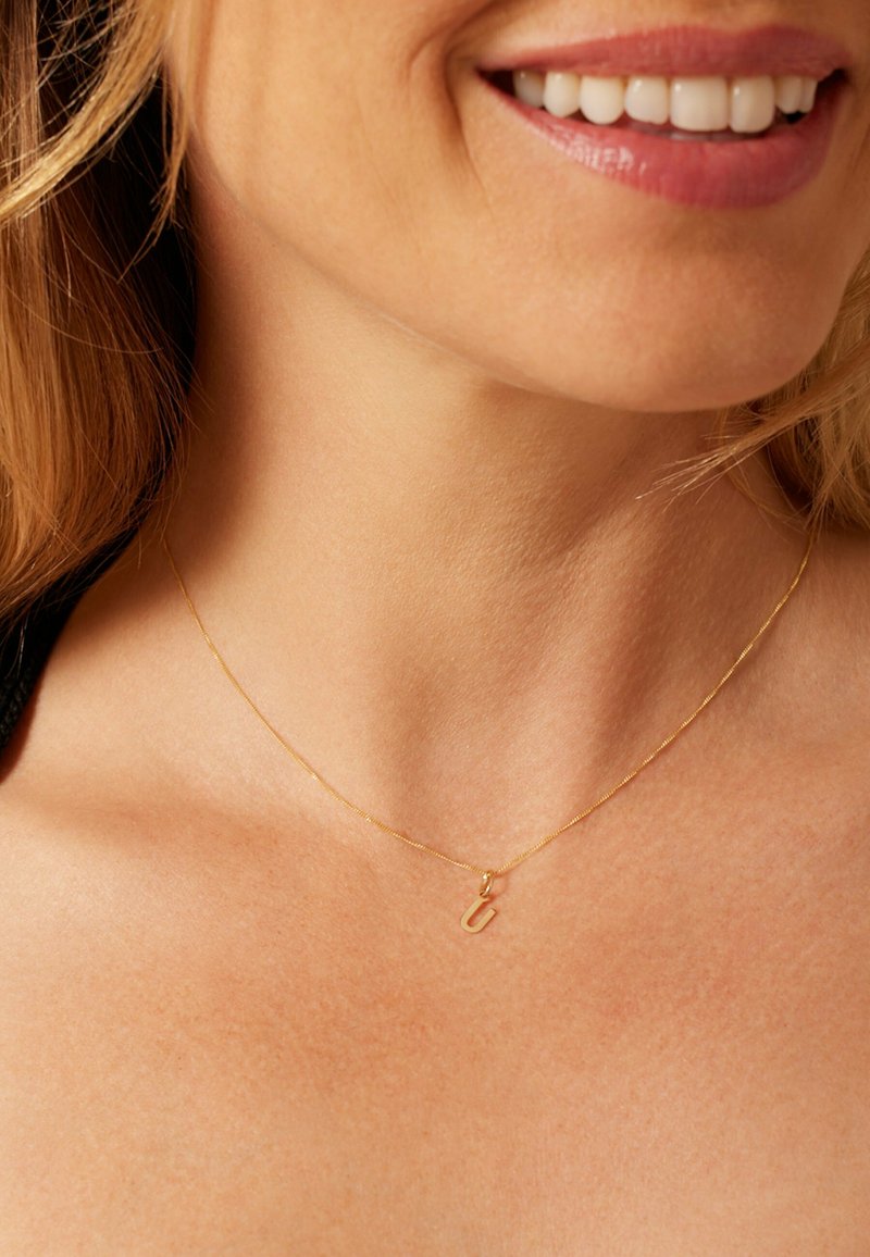 Gouden ketting met een delicaat hangertje in de vorm van een kleine letter. De ketting is dun en heeft een textuur, en ligt elegant op blote huid.