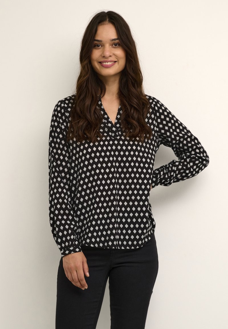 Blusa a maniche lunghe bianca e nera con motivo a diamante, colletto e chiusura frontale con bottoni, abbinata a jeans scuri. Tessuto con texture liscia.