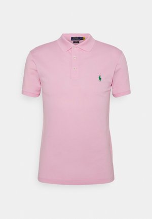 Jasnoróżowa koszulka polo z krótkim rękawem, kołnierzykiem, trzema guzikami i małym zielonym wyszywanym logo na lewej piersi.