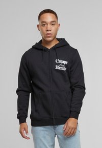 Sort zip-up hoodie med forlommer, snøre i huen og hvid tekstgrafik "Escape from Reality" på venstre bryst. Blød stofstruktur.