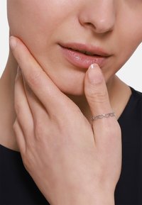 Anello d'argento con design a cuori intrecciati, indossato su un dito. Primo piano di una mano che tocca delicatamente il viso, mettendo in mostra la texture della pelle e i dettagli.