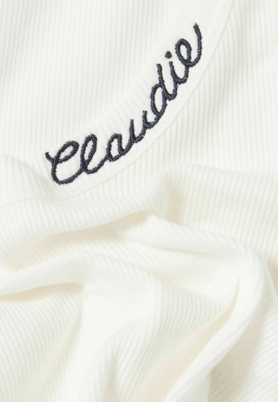 Tela blanca acanalada con texto en cursiva bordado en negro "Claudie" cerca de una costura o borde.