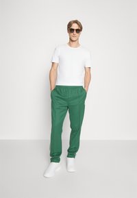 Green sweatpants with white side stripes, paired with a plain white t-shirt and white sneakers. The model wears dark sunglasses.