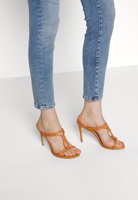 Sandalias de tacón alto naranja con un diseño de tiras, que cuentan con un lazo para el dedo del pie y un tacón delgado, combinadas con unos pantalones ajustados de mezclilla azul claro.
