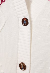 Biały dziergany kardigan z prążkowanymi brzegami i unikalnymi okrągłymi guzikami z ilustrowanymi postaciami w środku. Miękka faktura i casualowy design.