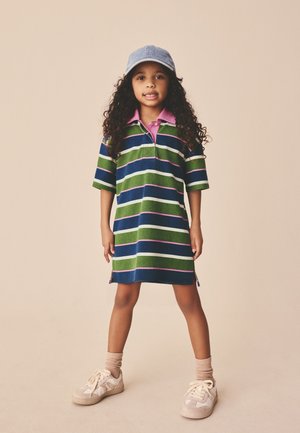 Joven niña con cabello largo y rizado que lleva un vestido polo a rayas, gorra azul claro, calcetas beige y zapatillas, parada con las piernas separadas.