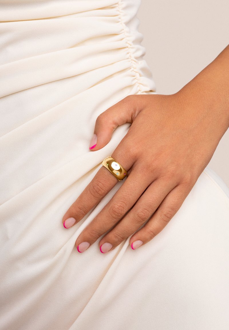 Goldring mit einem glatten, runden Design, das einen zentralen ovalen Edelstein und kleine Steine auf beiden Seiten zeigt. Die Hand ruht auf einem cremefarbenen Stoffhintergrund.