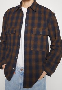 Brun och marinblå rutigt flanellskjorta med knäppning fram, två bröstfickor och långa ärmar. Bärs över en vit t-shirt och ljusa denimjeans.