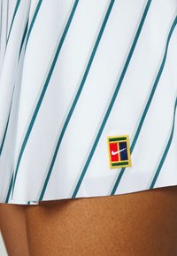 Falda blanca con rayas verticales en verde azulado, con un parche bordado del logo multicolor de Nike en el dobladillo. Textura de tela suave.