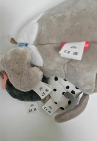 Peluche con tejido suave en gris y blanco, patrones de puntos y diversos acentos texturizados. Múltiples etiquetas de cuidado visibles en la parte inferior.