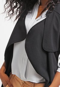Gilet nero in tessuto con frontale drappeggiato, finitura texture, indossato sopra una camicia button-up grigia chiara, abbinato a pantaloni beige. Capelli ricci visibili.