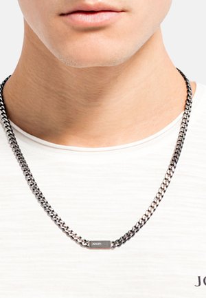 Zilverkleurige ketting met in elkaar grijpende schakels en een rechthoekig metalen label met de merknaam. Draag boven de halslijn van een witte blouse.