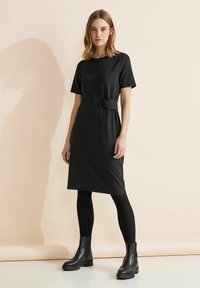 Robe noire à mi-mollet avec des manches courtes et un détail de nœud sur le côté à la taille. Associée à des leggings noirs et des bottes à semelles épaisses.