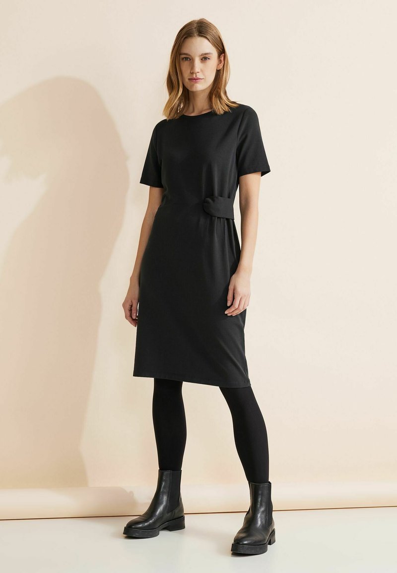 Robe noire à mi-mollet avec des manches courtes et un détail de nœud sur le côté à la taille. Associée à des leggings noirs et des bottes à semelles épaisses.