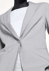 Blazer gris claro con solapas con muesca, cierre de botón simple y dos bolsillos frontales, hecho de un tejido suave con un corte entallado.