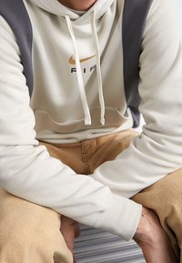 Ljusbeige hoodie med grå detaljer, med en framficka och dragsnören. Nike-logotypen i guld och svart. Tillverkad av bomull.