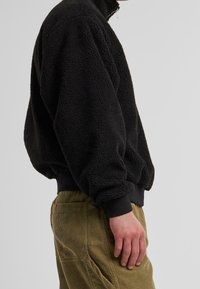 Sort fleece pullover med høj krave, tekstureret overflade, ribstrikkede håndled og kant. Båret med lysebrune каналbuks.