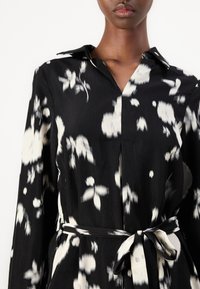 Μαύρο φόρεμα με λουλουδάτο μοτίβο που διαθέτει γιακά, μακριά μανίκια και ζώνη στη μέση με κόμπο. Το ύφασμα έχει μαλακή υφή και ρευστή πτώση.