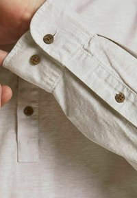 Ruka upravuje manžetu béžové košile s dlouhým rukávem hnědými knoflíky, přičemž je vidět podrobný šev a texturu látky.