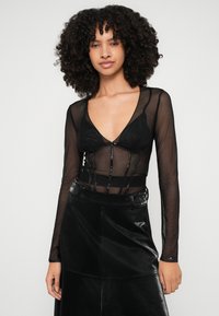 Fekete hálós, hosszú ujjú felső mély V-nyakkivágással és elülső csattal, fekete bralette-re rétegezve. Magas derekú fekete műbőr szoknyával párosítva.