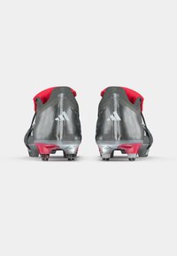 Botas de fútbol con una parte superior gris, acentos rojos y un acabado texturizado. Presentan un logo prominente y un diseño ligero y aerodinámico.