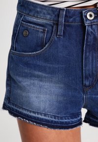 Tummansiniset farkkushortsit, joissa on revitty helma, etutasku, metallinappi ja vyölenkit. Shortsien pinnassa on lievää haalistumista ja ne tuntuvat sileiltä.