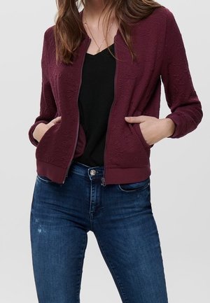 Vrouw draagt een bordeauxrode, getextureerde jas met ritssluiting over een zwart shirt, met handen in de zakken, gecombineerd met nauwsluitende blauwe spijkerbroek.