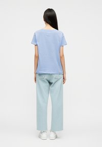 Ενήλικας με μακριά σκουρόχρωμα μαλλιά που φοράει ανοιχτό μπλε κοντομάνικο πουκάμισο, πλατύ παντελόνι τζιν ελαφρώς ξεβαμμένο και λευκά αθλητικά παπούτσια, στέκεται γυρισμένος προς τα πίσω.