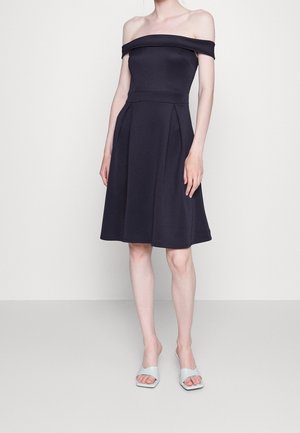 Kobieta w czarnej sukience za kolano, z odsłoniętymi ramionami oraz białych, otwartych sandałach na obcasie, stojąca na jednolitym tle.