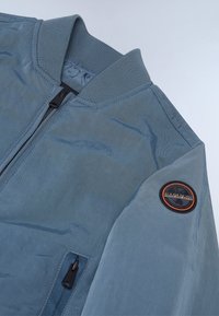 Lyseblå bomberjakke med ribbestrikket krage, svart glidelås og en sirkulær logo-patch på venstre erm. Glatt, strukturert stoff.