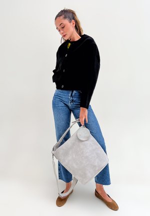 Femme avec une queue de cheval portant une veste noire, un jean bleu, des ballerines marron, tenant un grand sac à main gris clair avec une pochette ronde sur fond uni.
