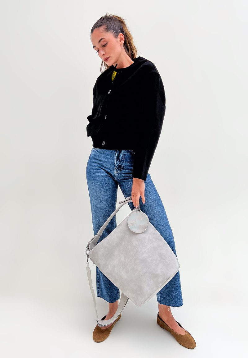 Femme avec une queue de cheval portant une veste noire, un jean bleu, des ballerines marron, tenant un grand sac à main gris clair avec une pochette ronde sur fond uni.