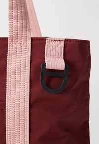 Mørkerød totebag med et blødt, teksturert stoff. Har lyserøde webbingstropper og en svart plastsløyfe.