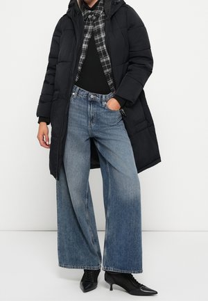 Personne portant un long manteau matelassé noir, une chemise à carreaux noir et blanc sur un haut noir, un jean bleu à jambes larges et des bottines noires à bouts pointus.