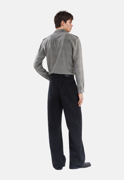 Langærmet, mønstret skjorte i grå og sort, parret med vide, teksturerede sorte corduroy bukser. Set fra ryggen.