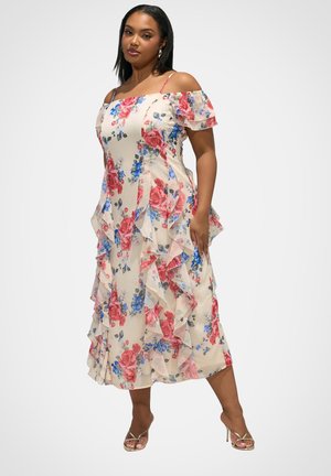 Vrouw die een crèmekleurige midijurk draagt met open schouders, roze en blauw bloemenprint, ruches, en metalen hoge hakken, staand tegen een witte achtergrond.