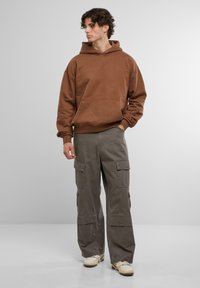 Hnědá, oversized mikina s přední kapsou; spárovaná se šedými cargo kalhotami s několika kapsami; neutrální tenisky dotvářejí celý outfit.