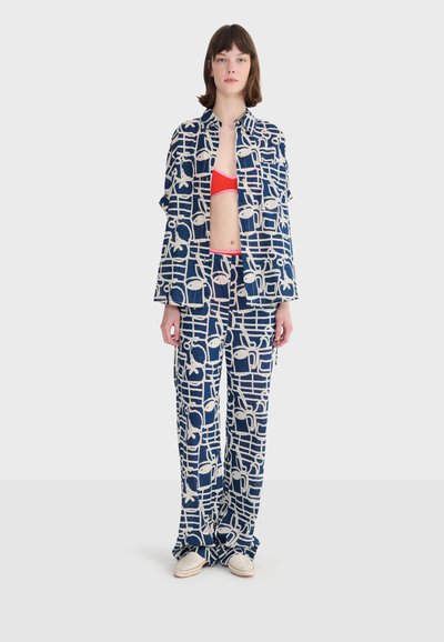 Młoda kobieta ubrana w luźną kurtkę i spodnie w niebiesko-biały wzór, czerwoną krótka bluzkę i lekkie plecione buty, stojąca na jednolitym tle.