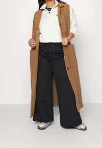 Lång brun ullväst med knäppning och fickor, buren över en vit zip-up sweatshirt och svarta vida byxor, kombinerad med silversandaler.