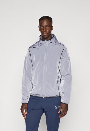 Lys grå, letvægts zip-up jakke med hætte, har sort piping langs sømmene og et logo på venstre brystområde.
