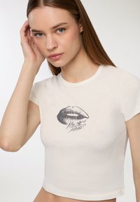 Kremowa, krótka koszulka z krótkim rękawem, na której znajduje się grafika ust oraz napis "Kiss Me Maybe?" wydrukowany na czarno. Miękka w dotyku.