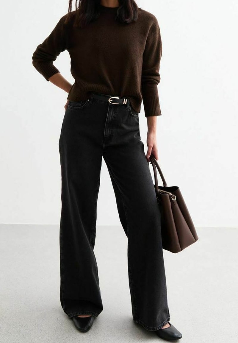 Personne portant un pull marron et un jean noir à jambes larges, tenant un sac à main marron dans la main droite, debout devant un fond blanc.