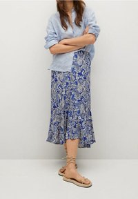 Camicia a righe blu e bianche con bottoni abbinata a una gonna blu a paisley. La gonna ha un orlo a campana. Indossata con sandali beige.