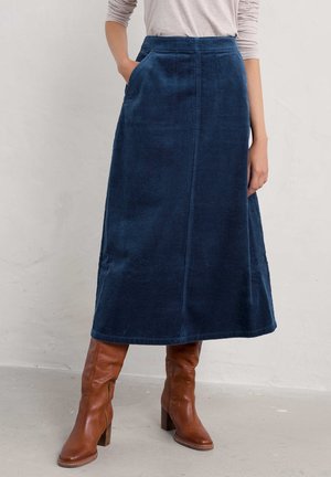 Femme portant une jupe bleu en velours côtelé longueur genou avec des poches et des bottes à talons en cuir marron mi-mollet, debout contre un mur uni.