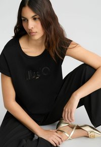 T-shirt nero a maniche corte con logo in rilievo "LIU･JO 1995" dalla texture lucida. Abbinato a pantaloni neri fluidi e scarpe dorate.