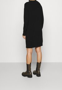 Persona con indosso un vestito in maglia nero a maniche lunghe e stivali alla caviglia chunky marrone scuro, in piedi con la schiena rivolta verso la fotocamera.