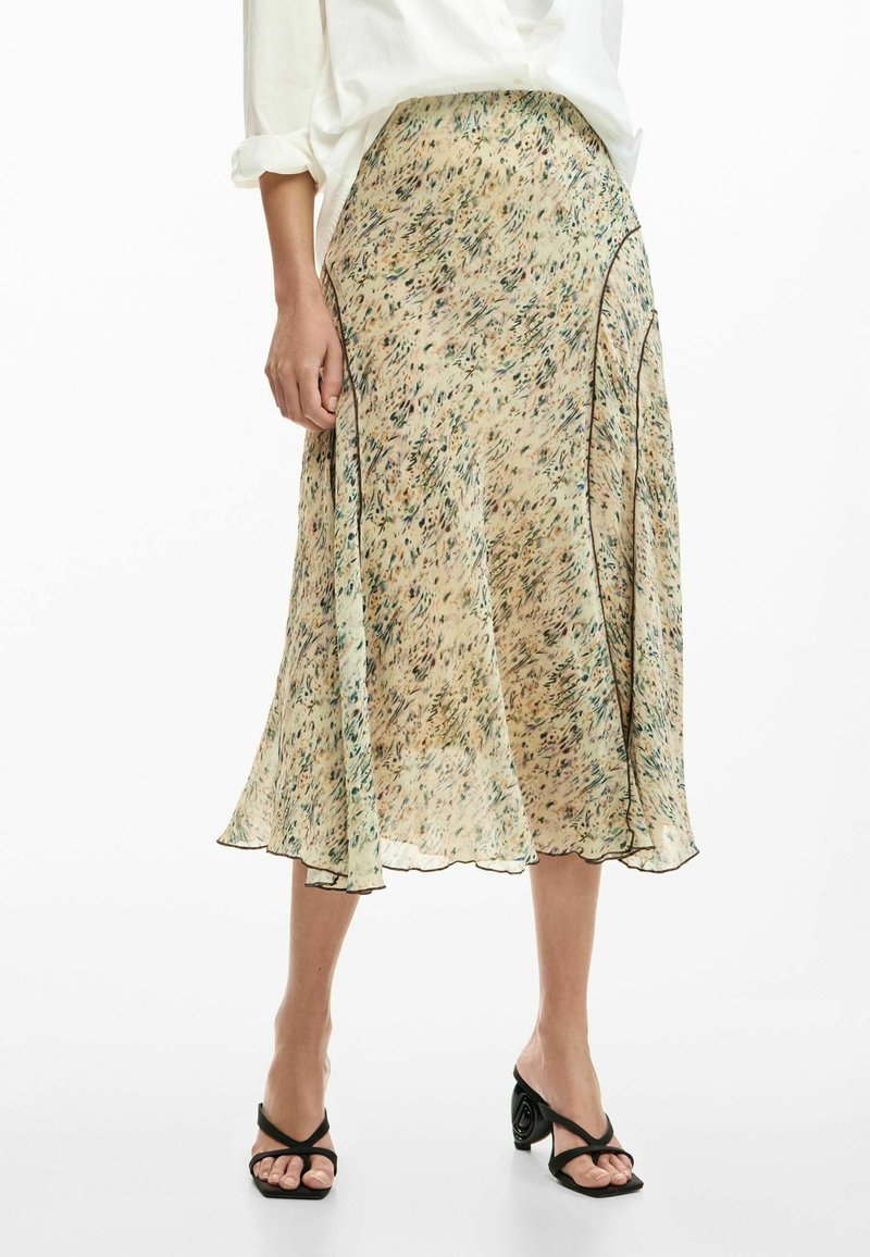 Donna che indossa una gonna midi beige a fiori con bordatura nera, camicetta bianca con maniche arrotolate e sandali neri con tacco, in piedi su sfondo bianco.