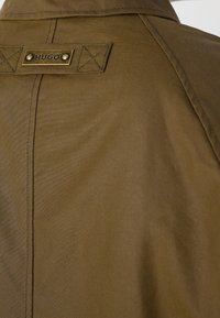 Gros plan du dos d'un manteau en tissu marron avec une plaque métallique cousue portant l'étiquette "HUGO" fixée près du col.